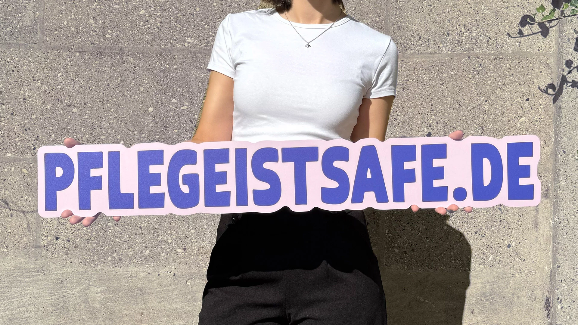 Person hält ein großes Schild mit der Aufschrift „PFLEGEISTSAFE.DE“ vor einer Betonwand.